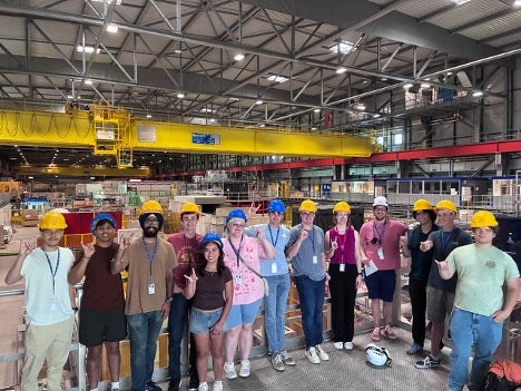 A group of students wearing hard hats pose for a photo in a warehouse, smiling and holding up the Hook 'Em, Horns hand sign.