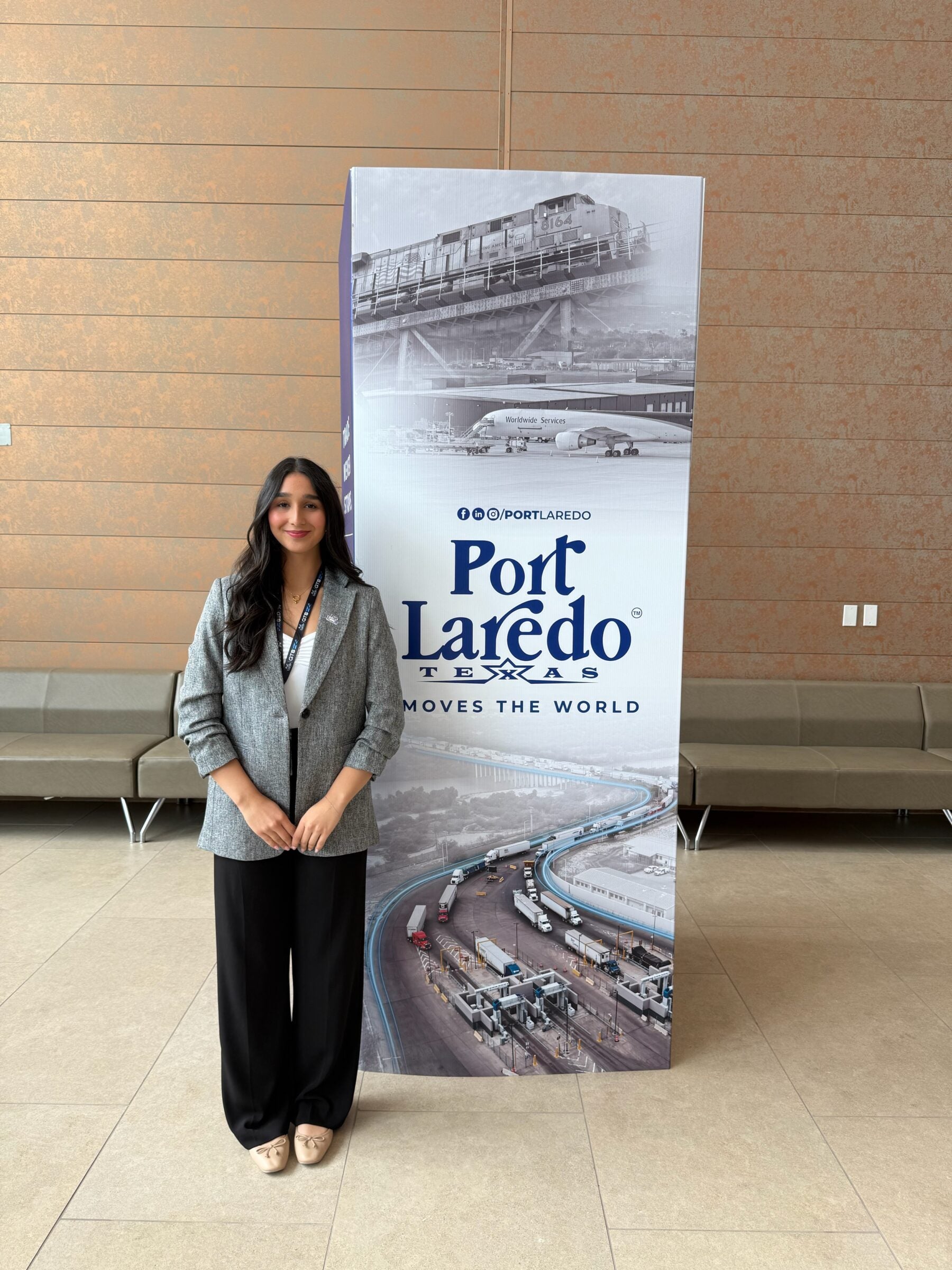 A girl wearing a grey blazer stands in front of a tall poster with "Port Laredo" written on it in blue font. 