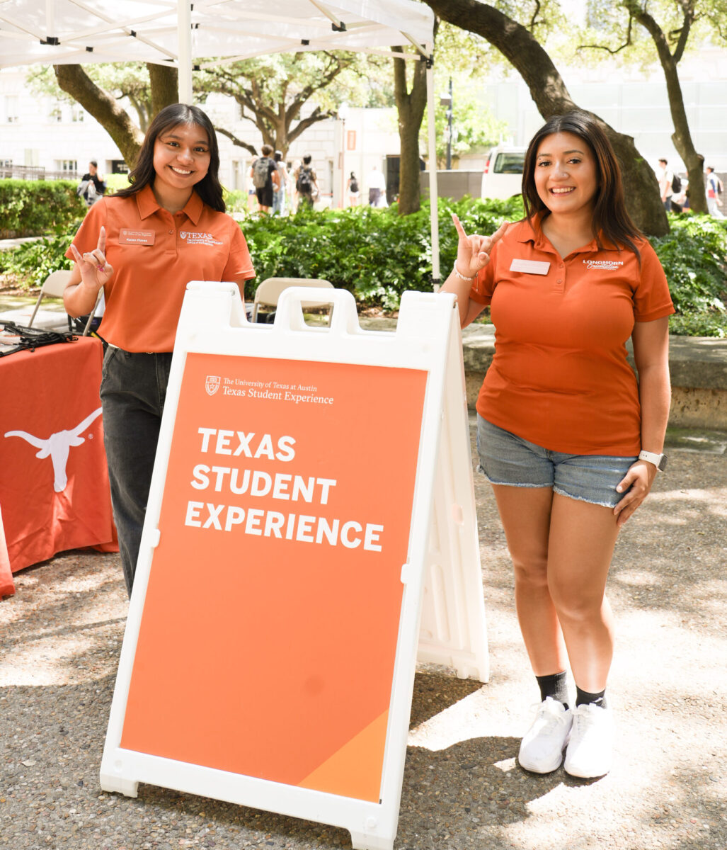 Two students in burnt orange pose outside under trees next to an orange sign