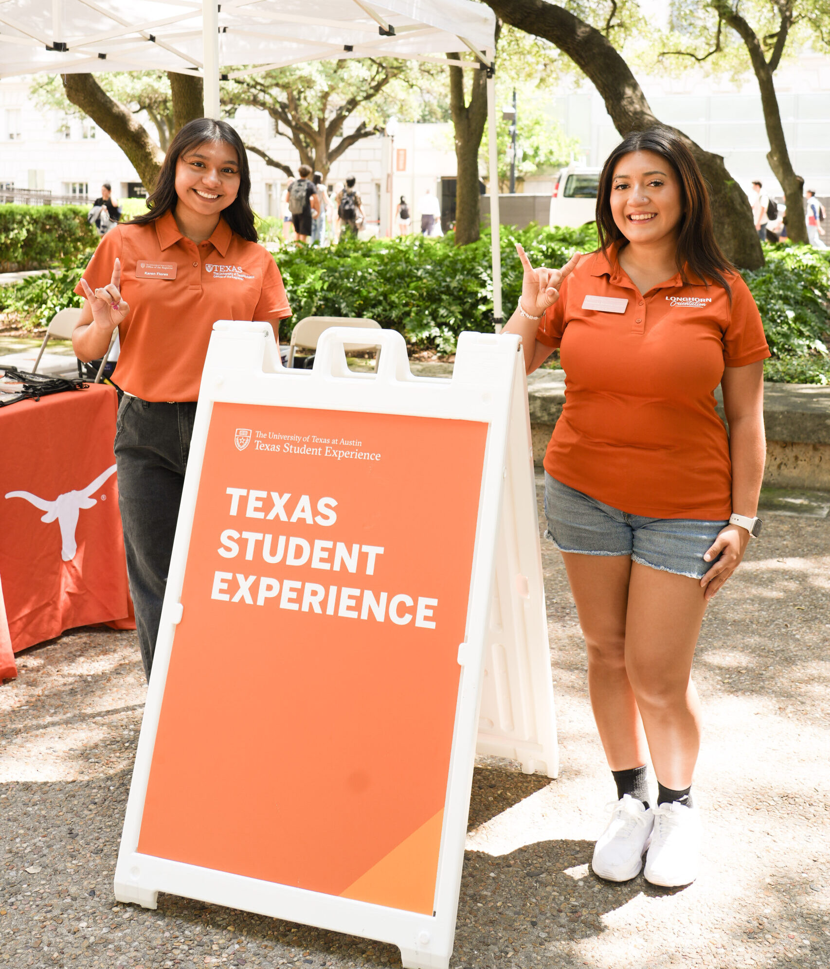 Two students in burnt orange pose outside under trees next to an orange sign