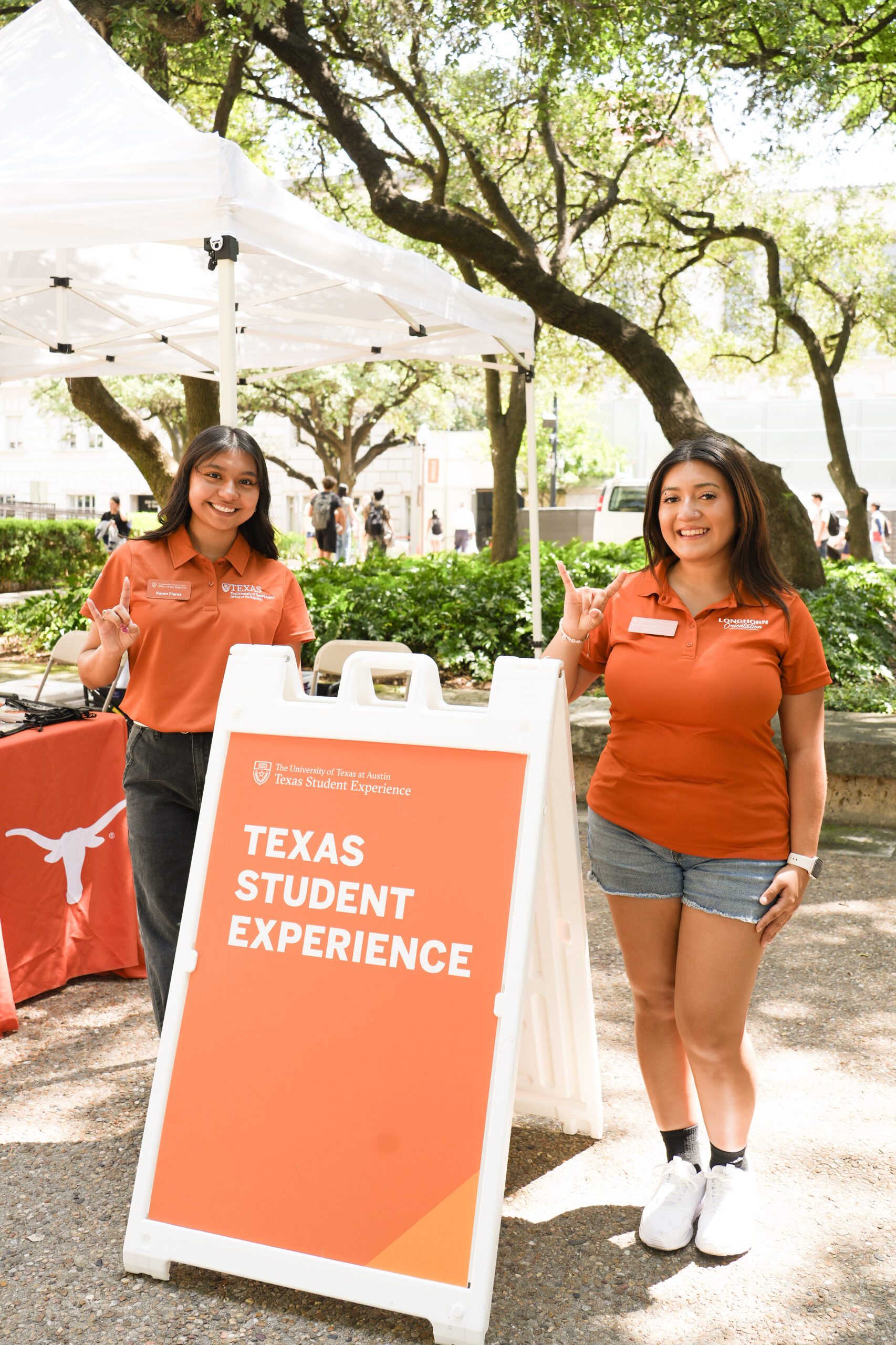 Two students in burnt orange pose outside under trees next to an orange sign