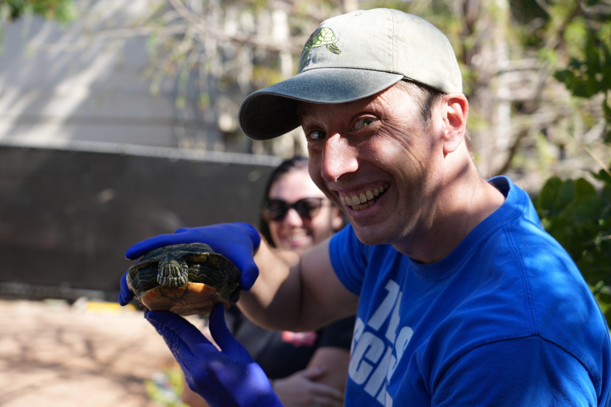 A man wearing a blue shirt and a ball cap holds a turtle up to the camera and smiles.