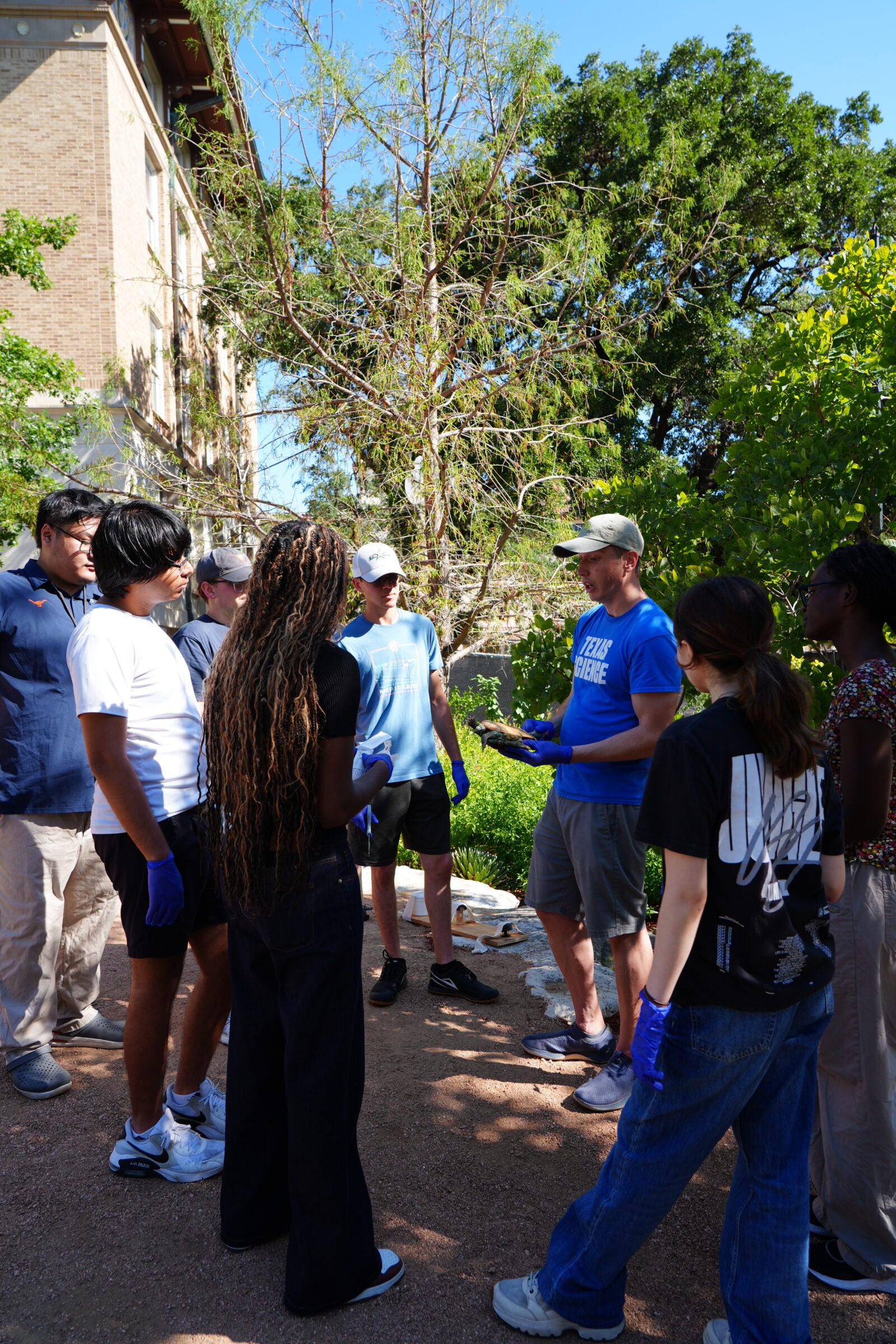 A group of students stand in a semi-circle around a man in a bright blue shirt. The man holds a turtle.