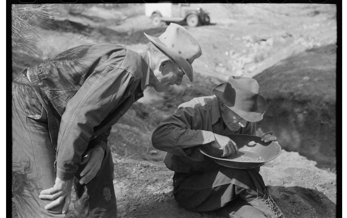 Gold prospector blowing away dirt to find the gold in his pan while a visiting prospector looks on. Pinos Altos, New Mexico. Credit: Library of Congress