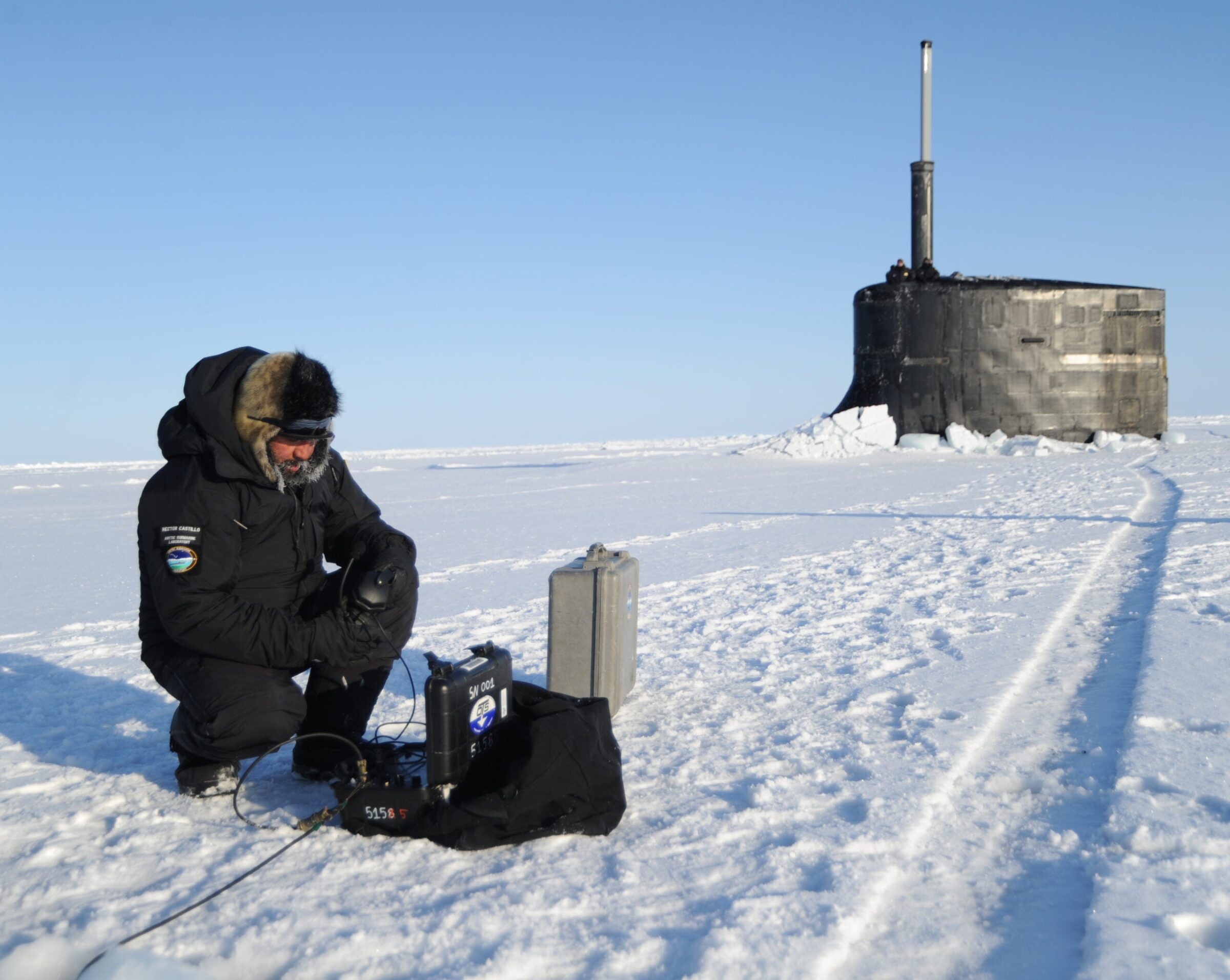 A researcher wearing a black parka works on a machine on snow-covered ground.