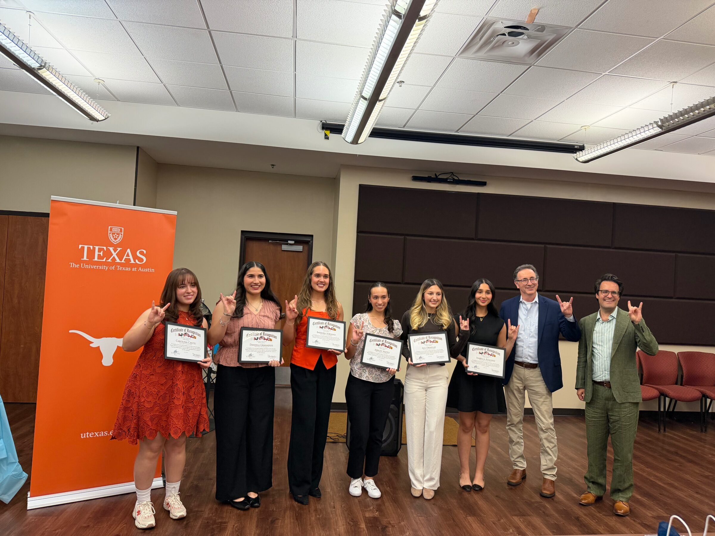 A group of six students stand in front of an orange University of Texas sign holding certificates. They are joined by two men in office wear.