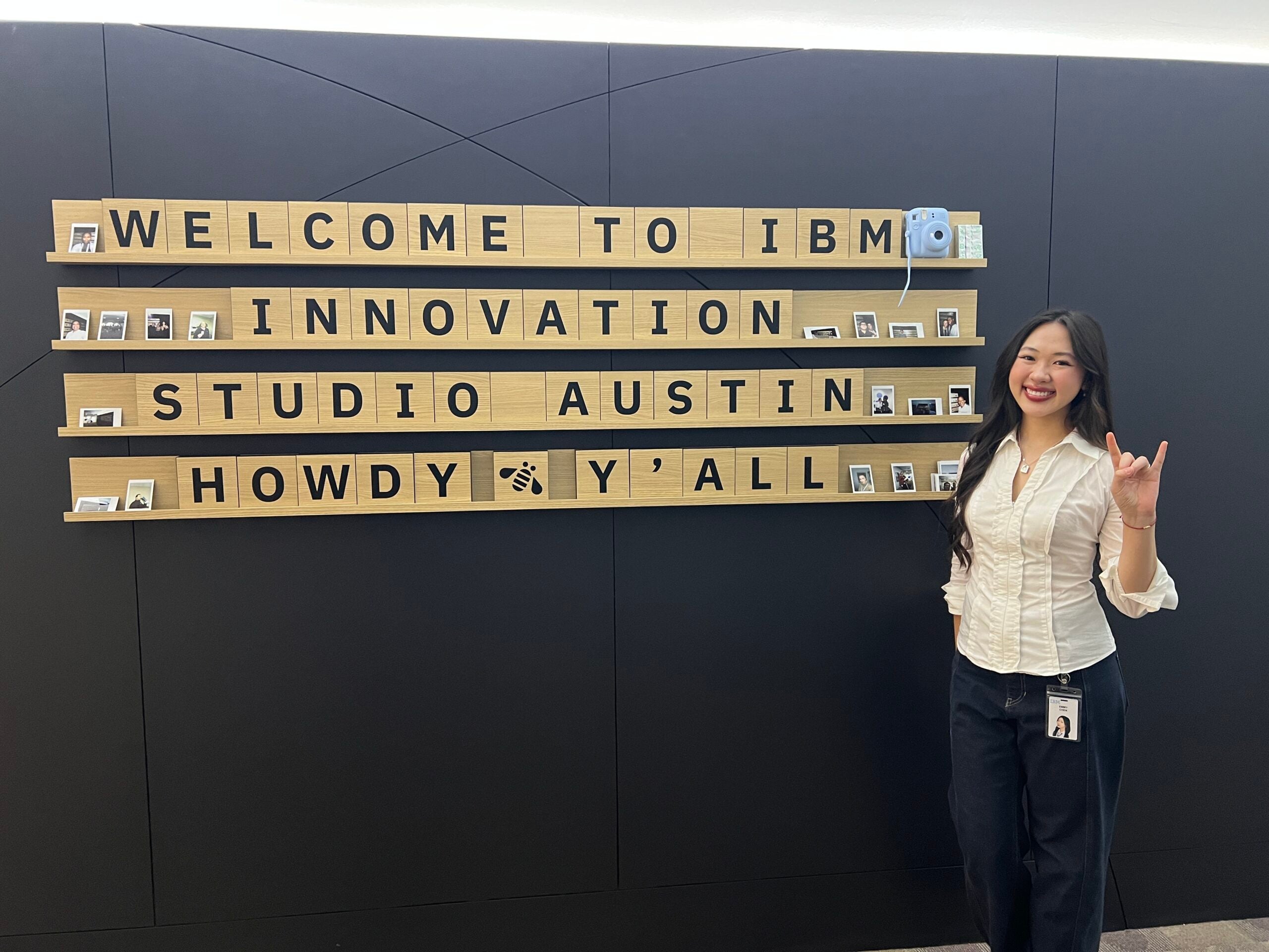 A woman poses in front of a display that reads "Welcome to IBM Innovation Studio Austin / Howdy Y'all"