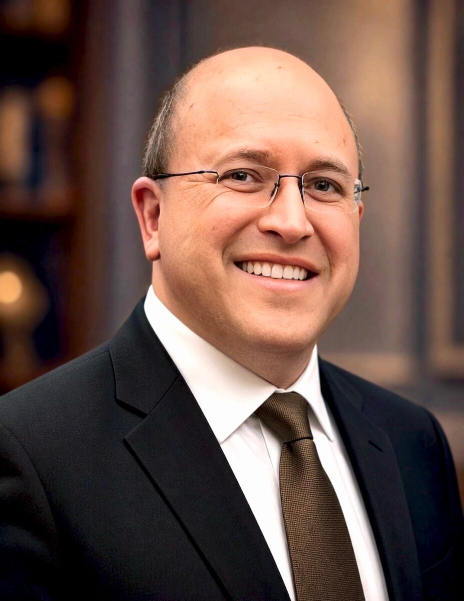 Man in suit, tie, and glasses poses outside in front of a building.