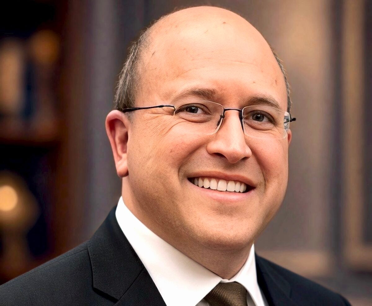Man in suit, tie, and glasses poses outside in front of a building.