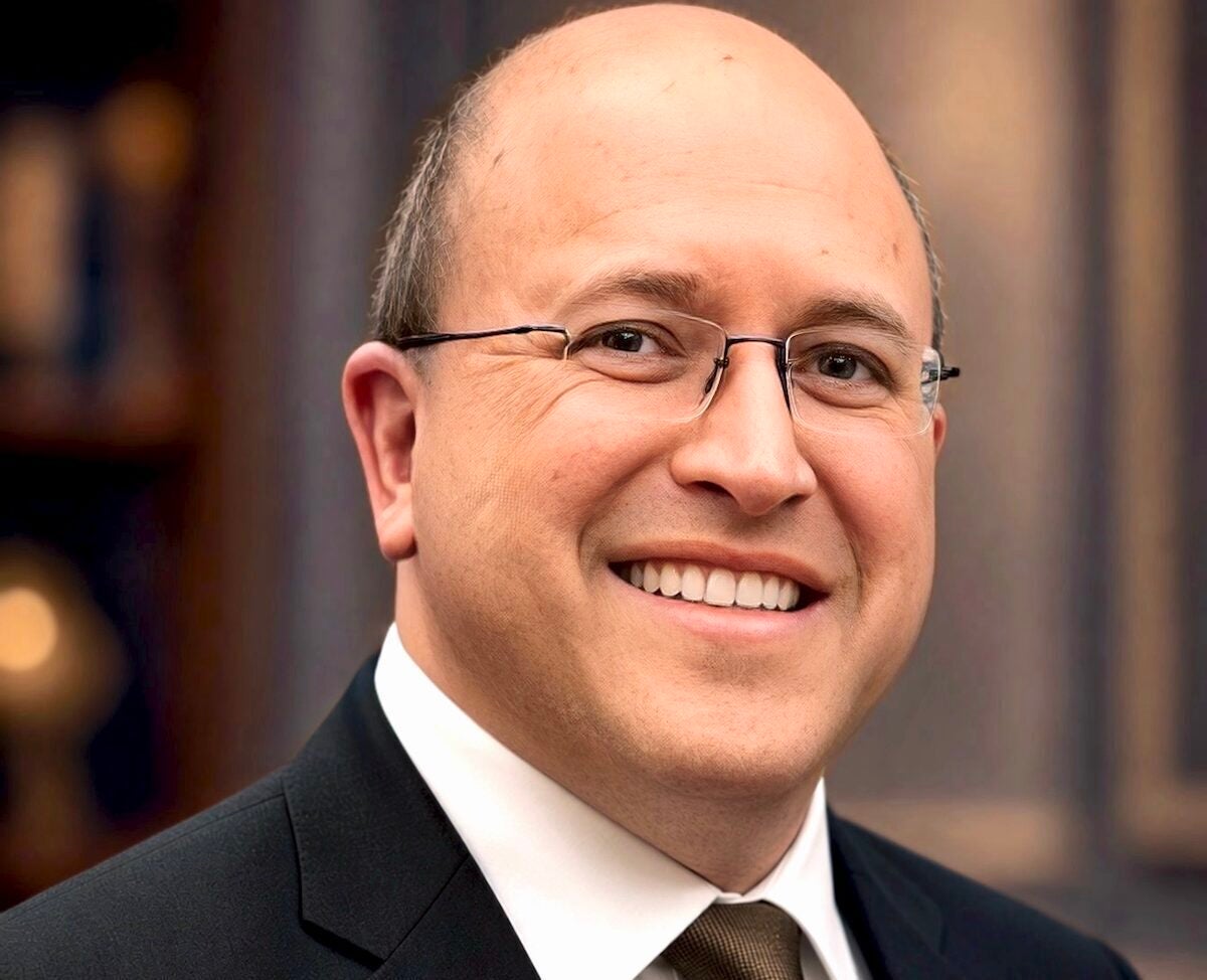 Man in suit, tie, and glasses poses outside in front of a building.