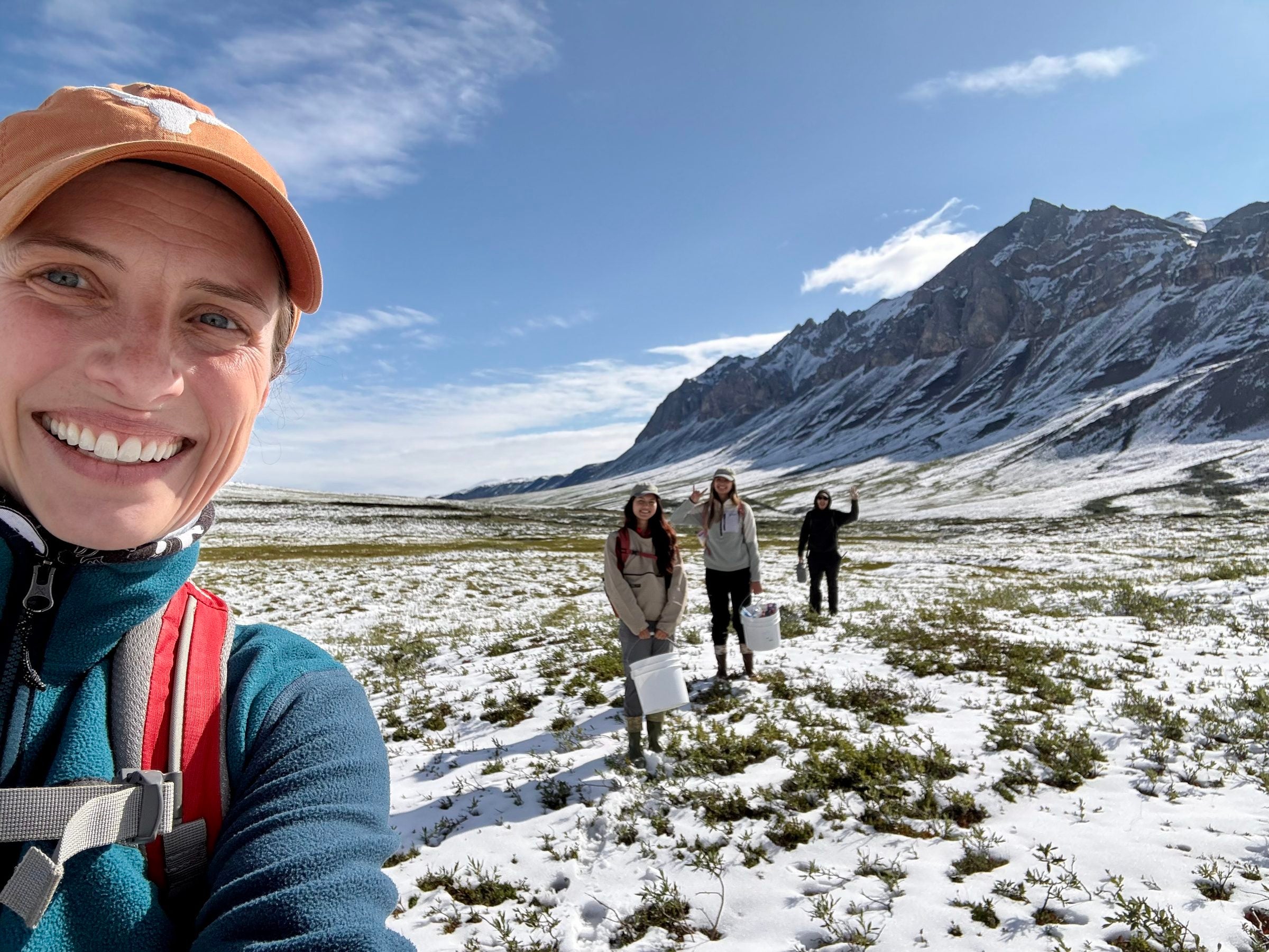 A smiling woman stands ahead of three people holding buckets in front of a snow covered field and mountain. They are posing for a selfie.