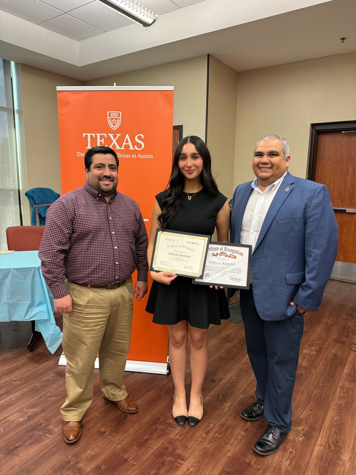 A girl in a black dress stands with two men and holds two paper certificates.