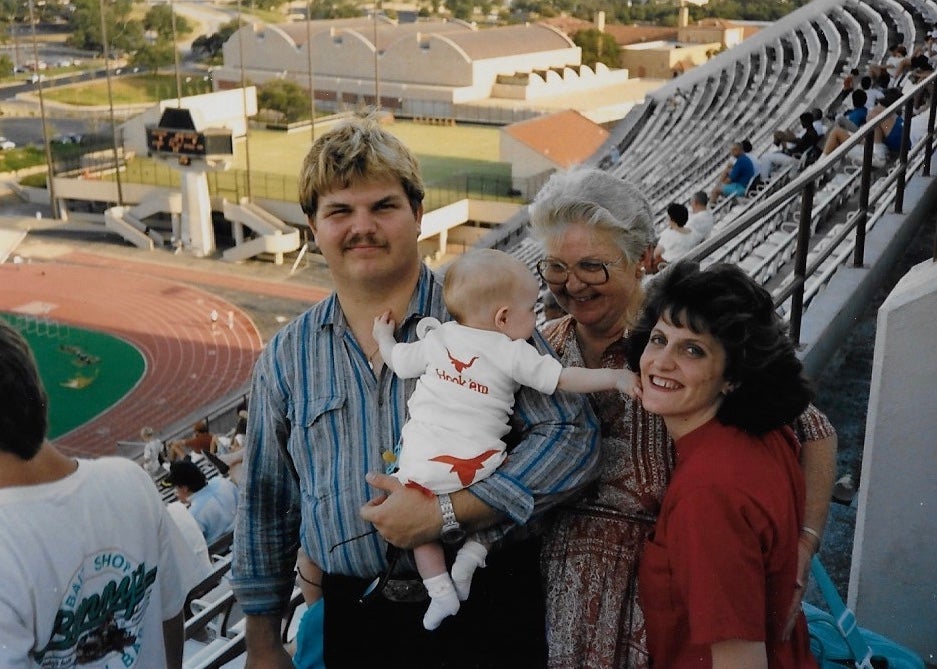 Three adults pose for a photo with a baby in the stands of Darrell K Royal-Texas Memorial Stadium in the 1990s.