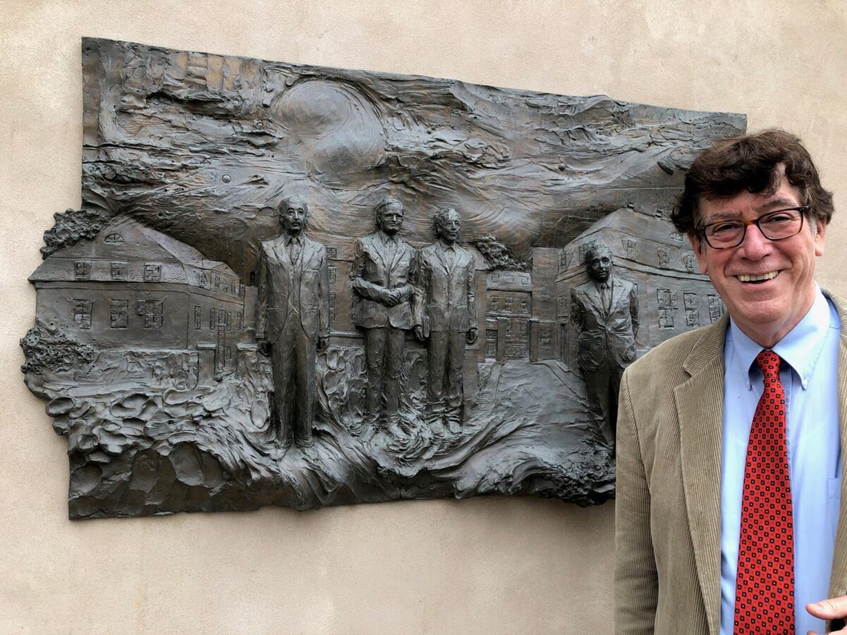 A man smiles for a photo in front of a bronze picture on a wall.