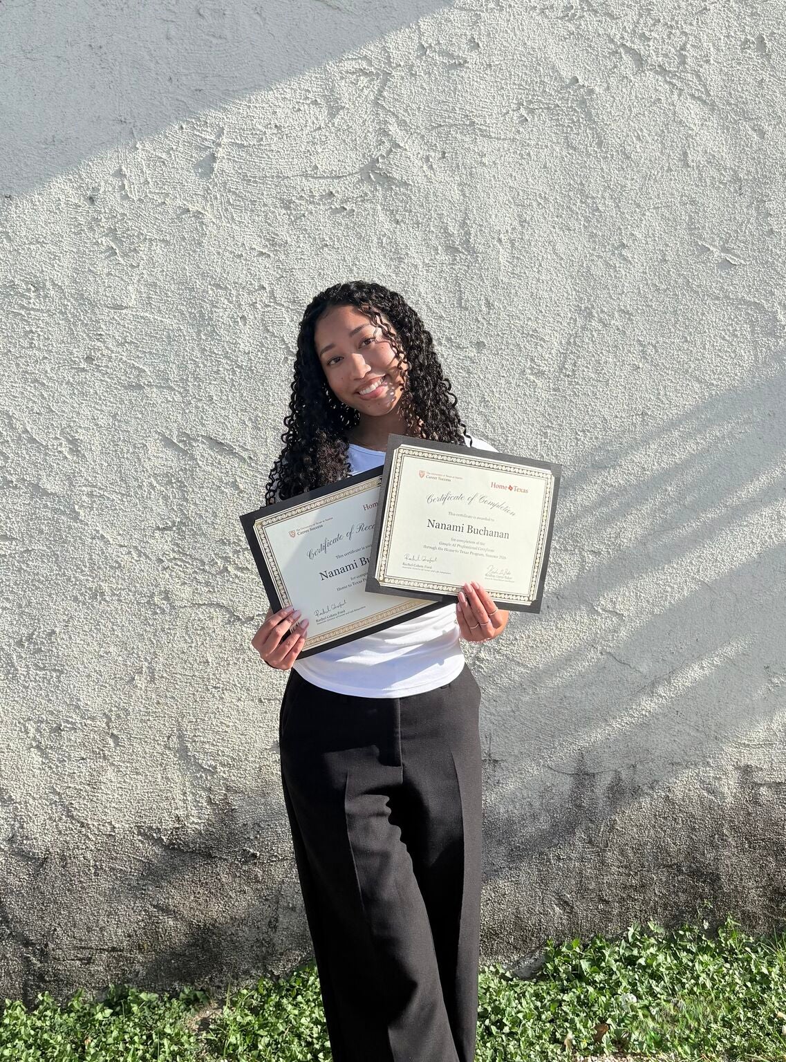 A girl stands in front of a white wall and holds two paper certificates. 