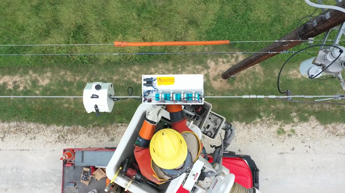 Utility worker in a vest and hard hat attaches a blue sensor to a power line.