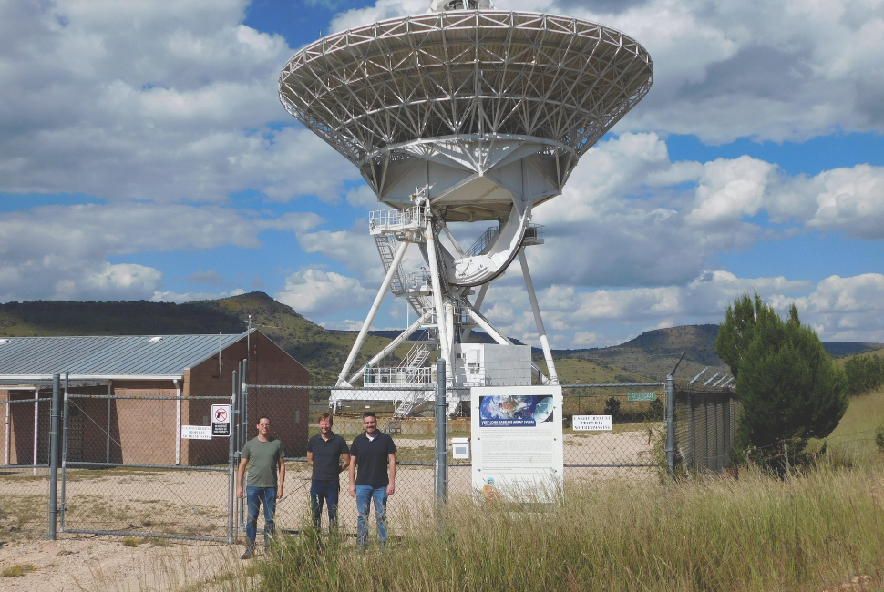 Three men pose for a photo in front of a radio antenna.