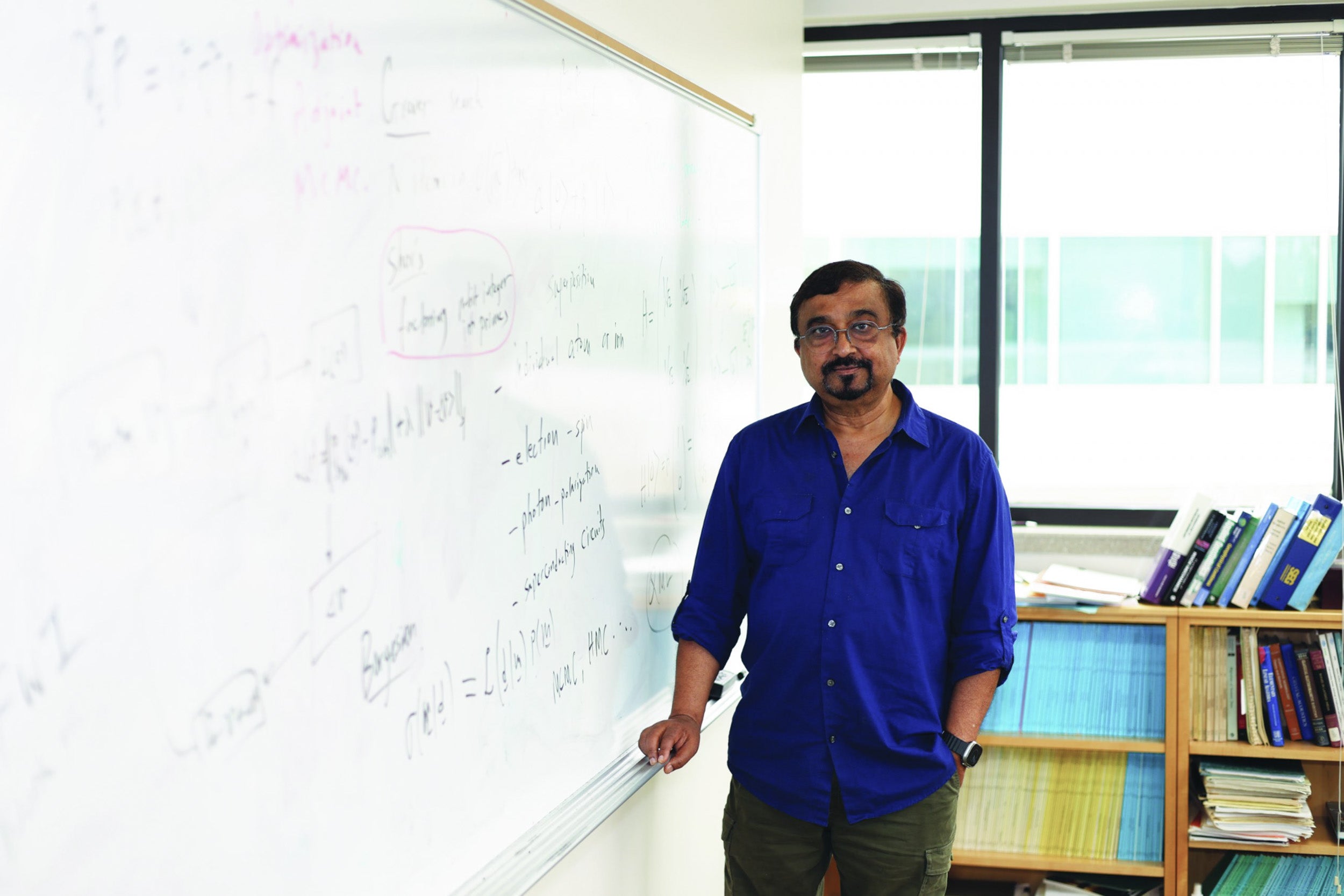 A man wearing a blue shirt and glasses poses for a photo next to a whiteboard.