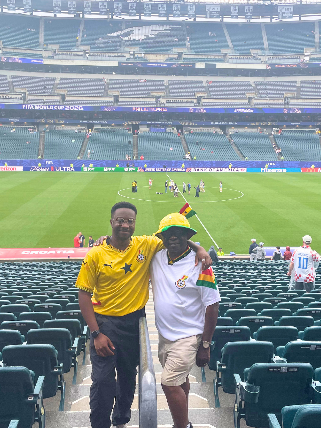 Two men pose for the photo in the stands of a soccer stadium.