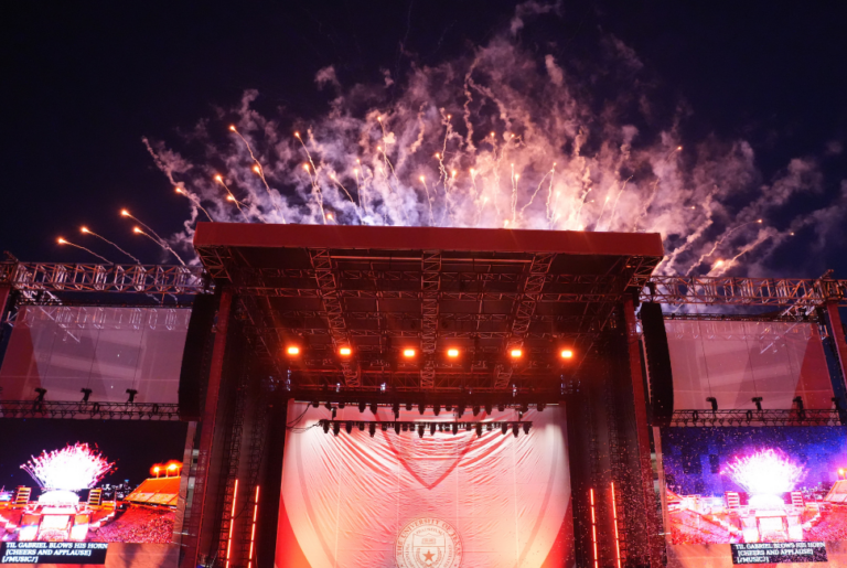 Fireworks light up the night sky behind a stage with two large screens on either side.