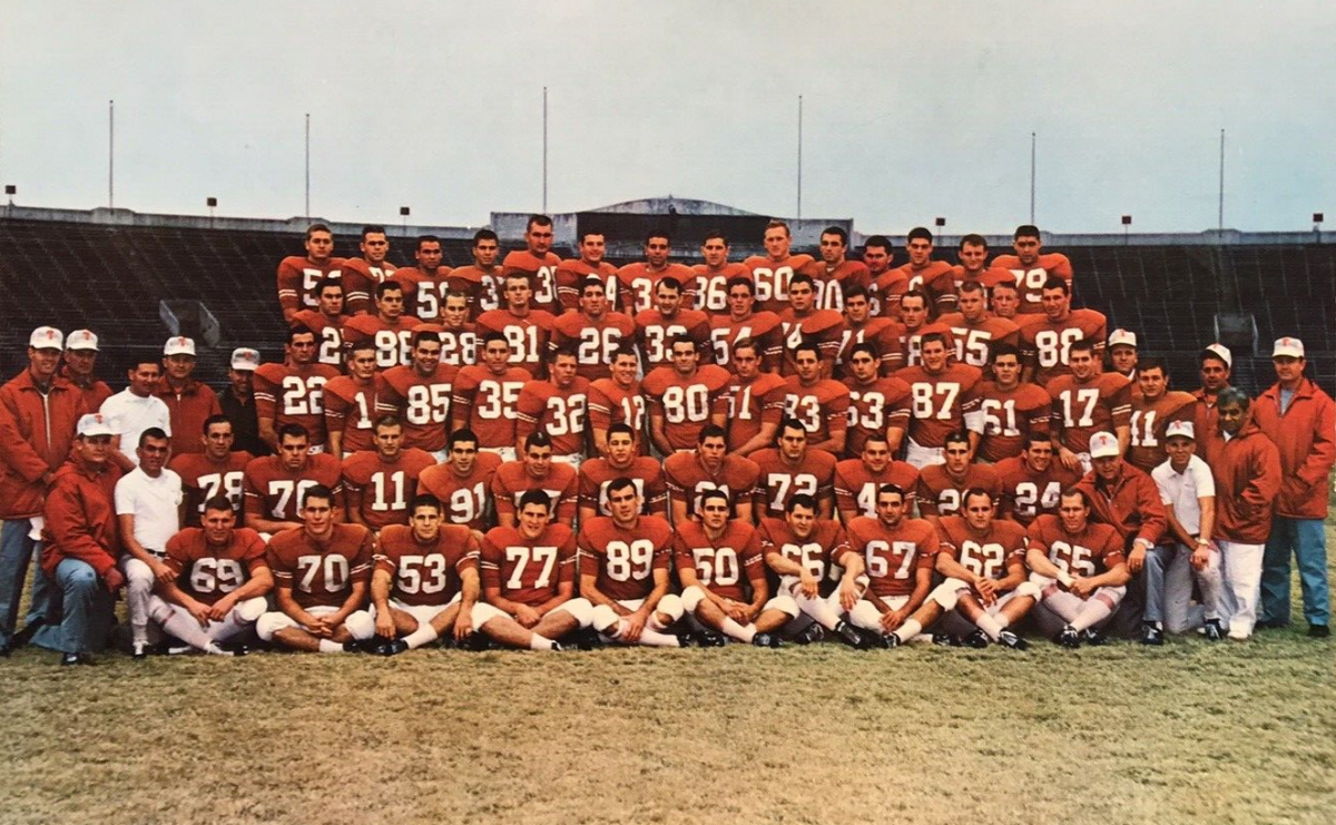 A football team wearing burnt orange uniforms sits together on the field with their coaches.