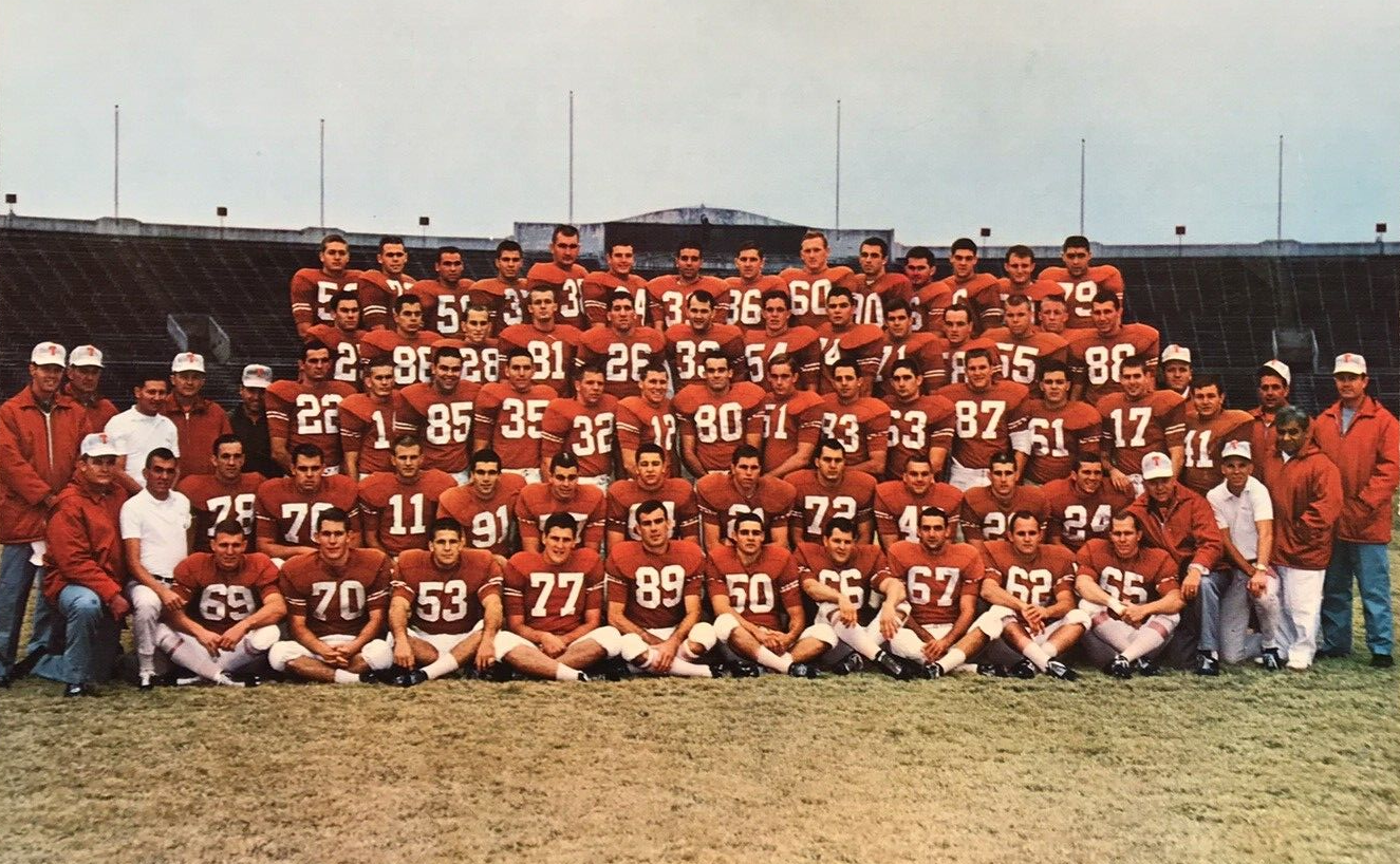A football team wearing burnt orange uniforms sits together on the field with their coaches.