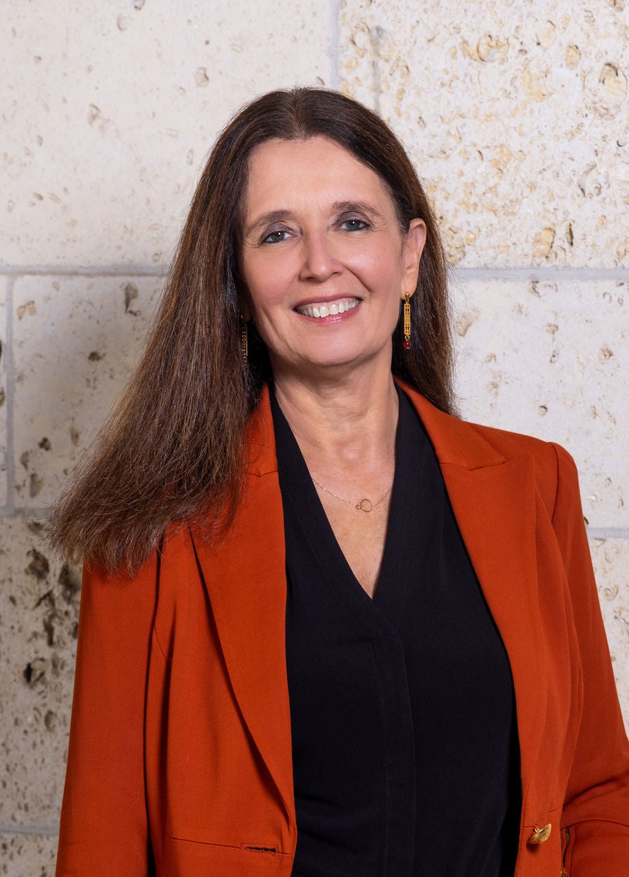 Woman in burnt orange jacket poses in front of a white stone wall.
