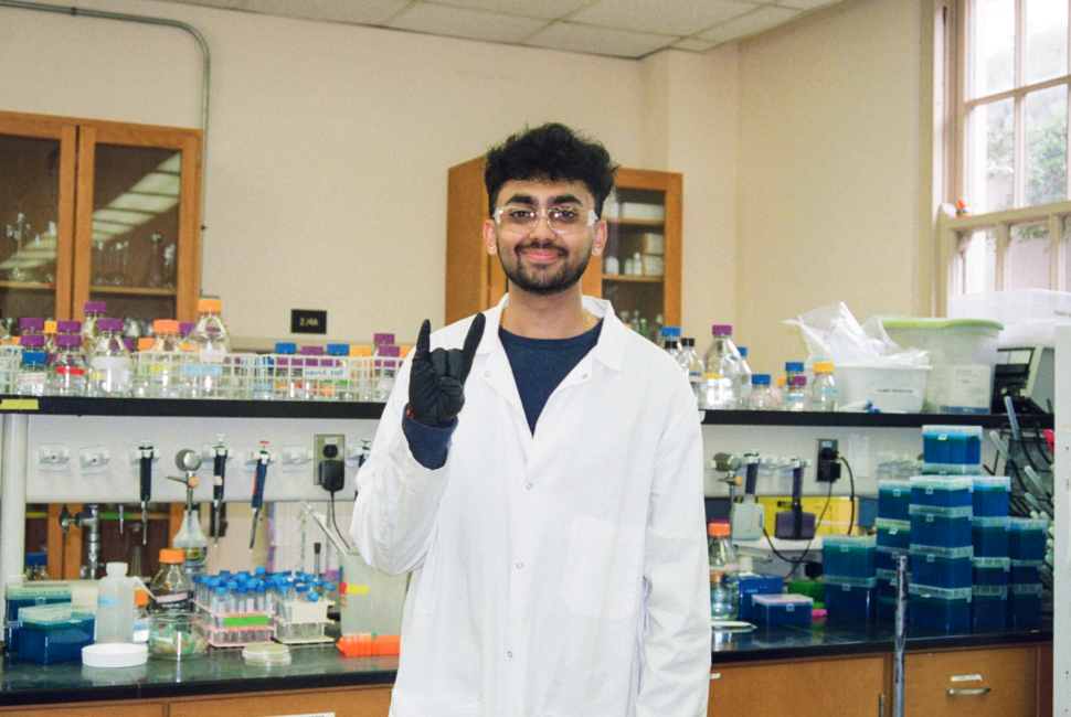 A student in a lab wears protective eyewear, a white coat and black gloves and holds up the Hook 'Em, Horns hand sign while smiling for a photo.