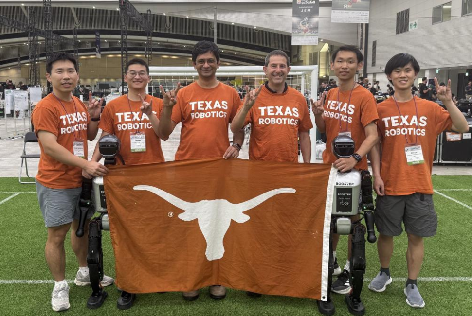 A group of people wearing burnt orange Texas Robotics T-shirts pose for a photo with a UT flag and two humanoid robots.