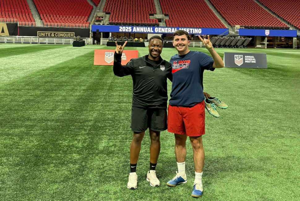 Two men pose for a photo on a soccer field, smiling and holding up the Hook 'Em, Horns hand sign.